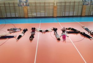 Zdjęcie pokazuje dzieci na sali gimnastycznej, które leżąc na podłodze układają napis ZIMA