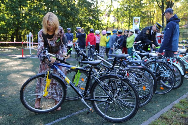 Egzamin na kartę rowerową w Zalesiu Górnym. Na zdjęciu wiceburmistrz Hanna Kułakowska- Michalak znakuje odblaskami rowery uczestników egzaminu. W tle uczniowie przygotowujący się do egzaminu.