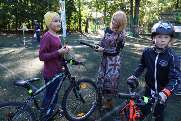 Egzamin na kartę rowerową w Zalesiu Górnym. Na zdjęciu wiceburmistrz Hanna Kułakowska-Michalak wręcza uczniowi odblask do roweru. W tle inni uczestnicy egzaminu.