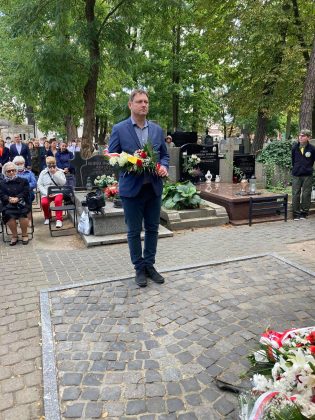elegancko ubrany mężczyzna z kwiatami składa hołd pod pomnikiem Katyńskim