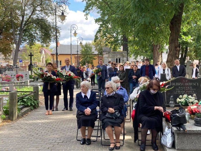 widok na uczestników uroczystości. Część osób stoi pod drzewami, część zajęła miejsca siedzące.
