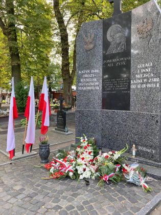 Pomnik Katyński z szarego kamienia, u jego szczytu napis "pomordowani ale żyją...". pod pomnikiem złożone wieńce, a obok trzy biało czerwone flagi.
