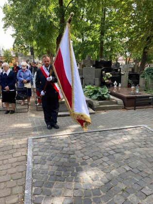 mężczyzna przepasany biało-czerwona wstęgą staje pod pomnikiem katyńskim ze sztandarem, w tle uczestnicy uroczystości i cmentarz
