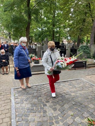 dwie starsze kobiety idą z kwiatami w stronę pomnika. Ich twarze zakrywają maseczki ochronne.