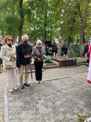 trójka starszych osób, dwie kobiety i mężczyzna stoją pod pomnikiem. Ich twarze zakrywają maseczki ochronne.
