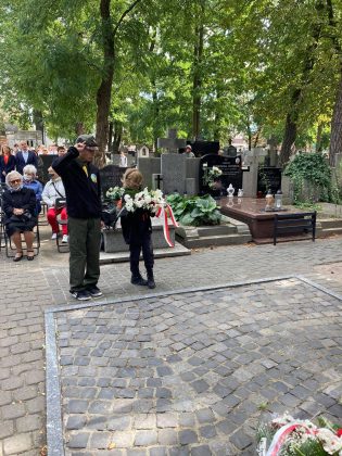 Mała dziewczynka z bukietem białych kwiatów i starszy od niej harcerz w mundurze oddają hołd pod pomnikiem.