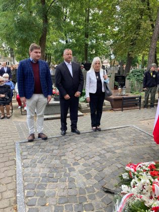 trzy osobowa delegacja - jedna kobieta i dwóch mężczyzn - stoją pod Pomnikiem Katyńskim, za ich plecami uczestnicy uroczystości.