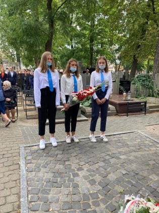 Młodzież szkolna - trzy młode dziewczyny w odświętnych strojach oddają hołd pod pomnikiem.Młodzi ludzie maja na sobie białe koszule i niebieskie krawaty.