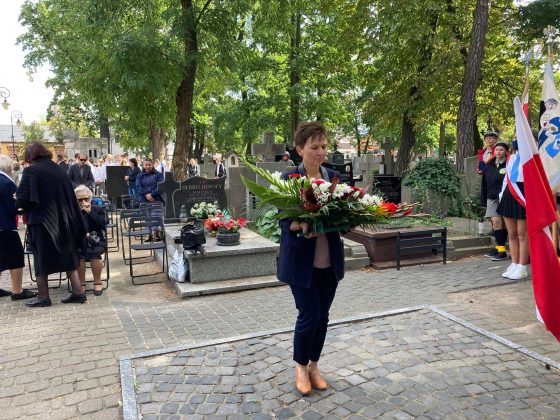 Członek Zarządu Powiatu Piaseczyńskiego Ewa Lubianiec składa pod pomnikiem wiązankę kwiatów w biało-czerwonych barwach. W tle uczestnicy uroczystości.