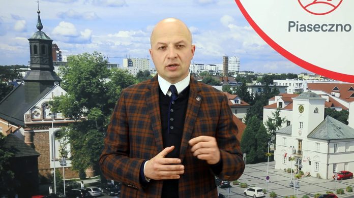 Na zdjęciu Daniel Putkiewicz - Burmistrz Miasta i Gminy Piaseczno, który stoi na tle banera reklamowego ze zdjęciem Ratusza i kościoła Św. Anny w Piasecznie