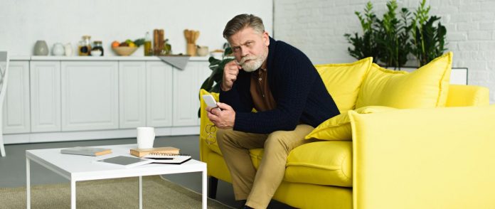 Ilustracja. Na zdjęciu Pan siedzi na żółtej kanapie i trzyma telefon w ręku. Zasady odbywania kwarantanny i izolacji obowiązujące od 2 września 2020 r.