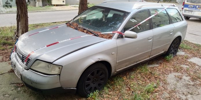 Ilustracja. Wraki pojazdów na terenie gminy Piaseczno. Na zdjęciu wrak srebrnego, opuszczonego samochodu oklejony taśmą straży miejskiej.