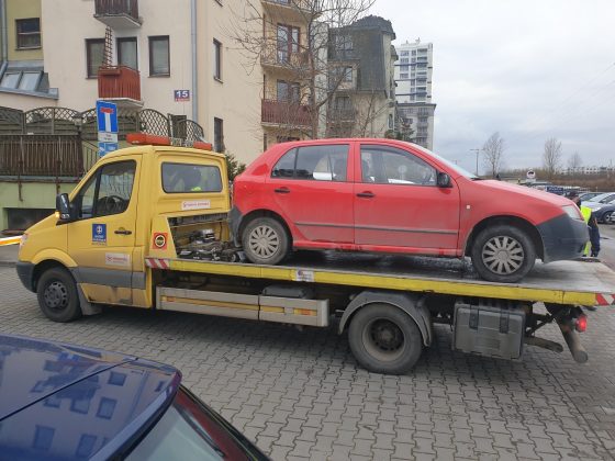 Ilustracja. Wraki pojazdów na terenie gminy Piaseczno. Na zdjęciu samochód laweta z odholowanym czerwonym samochodem. W tle bloki.