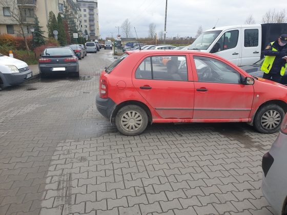 Ilustracja. Wraki pojazdów na terenie gminy Piaseczno. Na zdjęciu czerwony samochód odholowywany przez Straż Miejską w Piasecznie. W tle budynki i inne zaparkowane samochody.