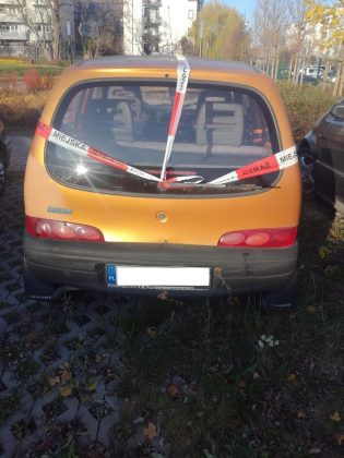 Ilustracja. Wraki pojazdów na terenie gminy Piaseczno. Na zdjęciu pomarańczowy samochód oklejony taśmą straży miejskiej.