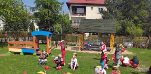 Ilustracja. Grupa dzieci na dworze, na trawniku bierze udział w zawodach sportowych. Dzieci kucają, na głowie trzymają kolorowe woreczki. Pomiędzy nimi leżą czerwone piłki. W tle widać plac zabaw i budynek.