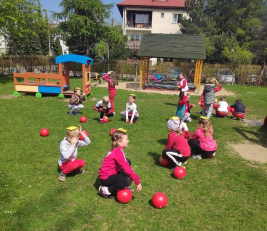 Ilustracja. Grupa dzieci na dworze, na trawniku bierze udział w zawodach sportowych. Dzieci kucają, na głowie trzymają kolorowe woreczki. Pomiędzy nimi leżą czerwone piłki. W tle widać plac zabaw i budynek.