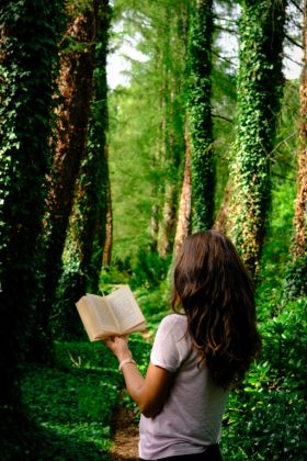 Kobieta odwrócona tyłem do fotografującego czyta księżkę. Otaczają ją zielone drzewa porośnięte bluszczem.