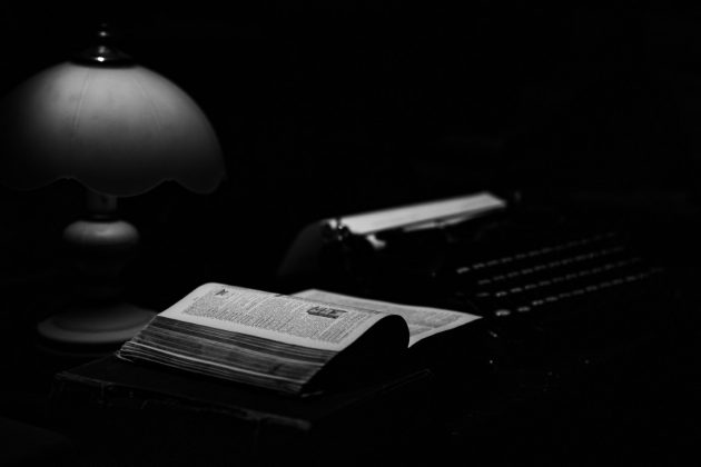 Czarnobiała fotografia przedstawiająca otwarta książkę. W tle maszyna do pisania.
