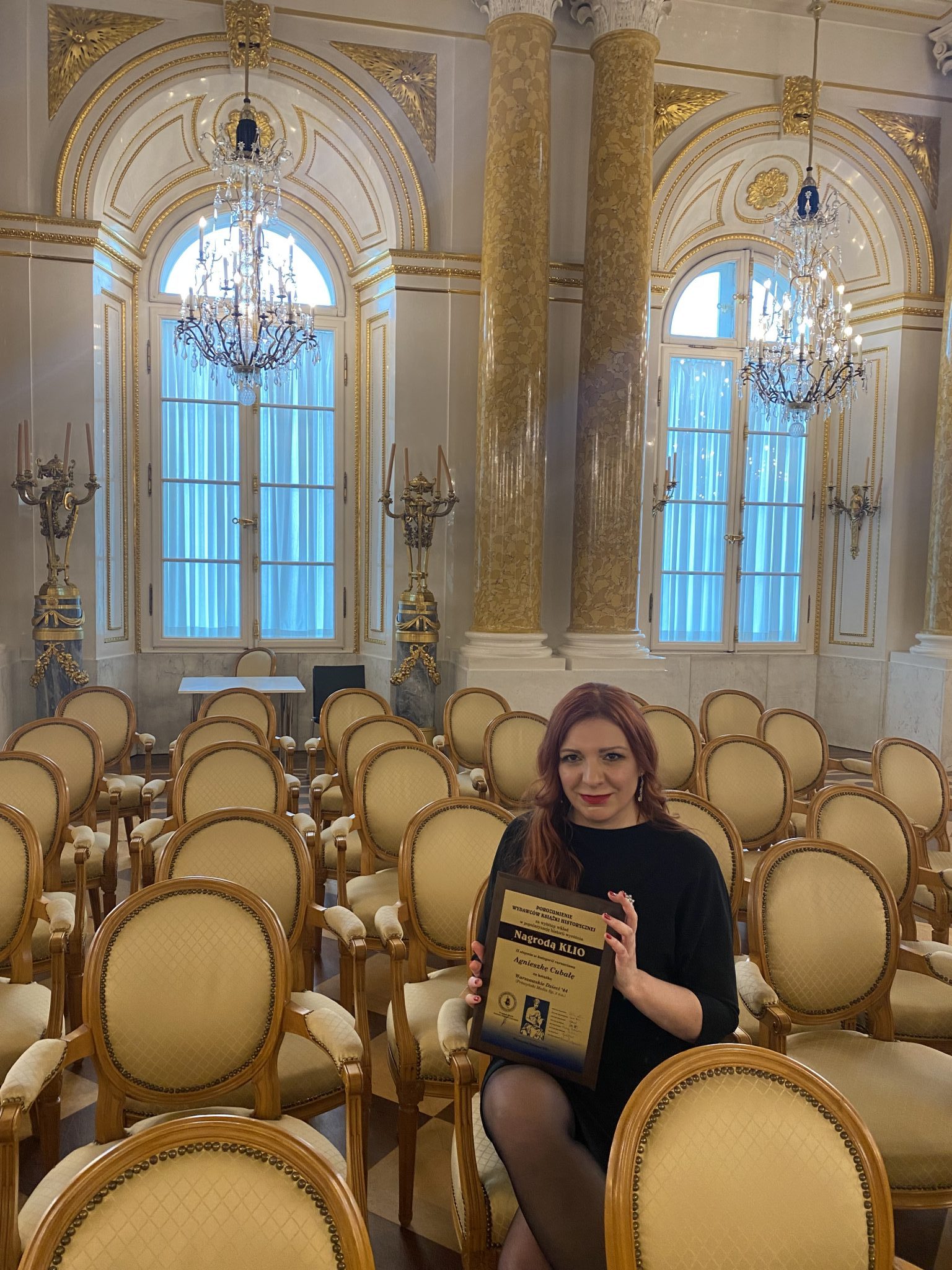 Agnieszka Cubała laureatką prestiżowej nagrody KLIO. Na zdjęciu autorka w sali Zamku królewskiego, siedzi na krześle i prezentuje dyplom.