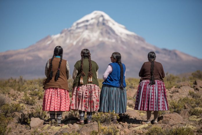 Pegaz na biegunach. Siła Spódnicy - Cholitas z Boliwii. Na zdjęciu Boliwijki w tradycyjnych strojach, odwrócone plecami do obiektywu, a w tle ośnieżony szczyt góry.