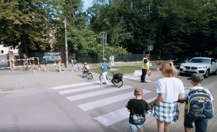 bezpieczna droga do szkoły. Na zdjęciu rodzice i dzieci przechodzacy przez przejście, strażnik miejski, samochody.