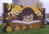 Mural Biblioteki. Namalowana twarz w okularach wystająca zza ksiązki, wokół liście dębu, kolorystyka jesienna.