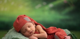 Na zdjęciu noworodek w czerwonej czapeczce i ubranku na zielonym tle.