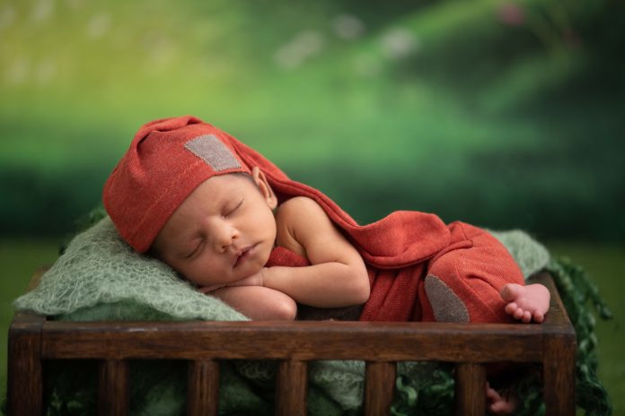 Na zdjęciu noworodek w czerwonej czapeczce i ubranku na zielonym tle.
