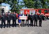 Dotacje Mazowsze straż pożarna . Na zdjęciu strażacy, dwóch mężczyzn bez mundurów i jedna kobieta. W tle wozy strażackie.