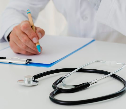 słuchawki lekarskie i dłoń z długopisem oparta o kartkę na biurku