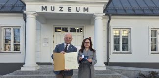 Burmistrz i dyrektor muzeum pozują pod budynkiem muzeum z dyplomem i medalem