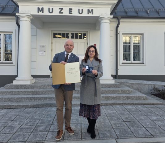 Burmistrz i dyrektor muzeum pozują pod budynkiem muzeum z dyplomem i medalem