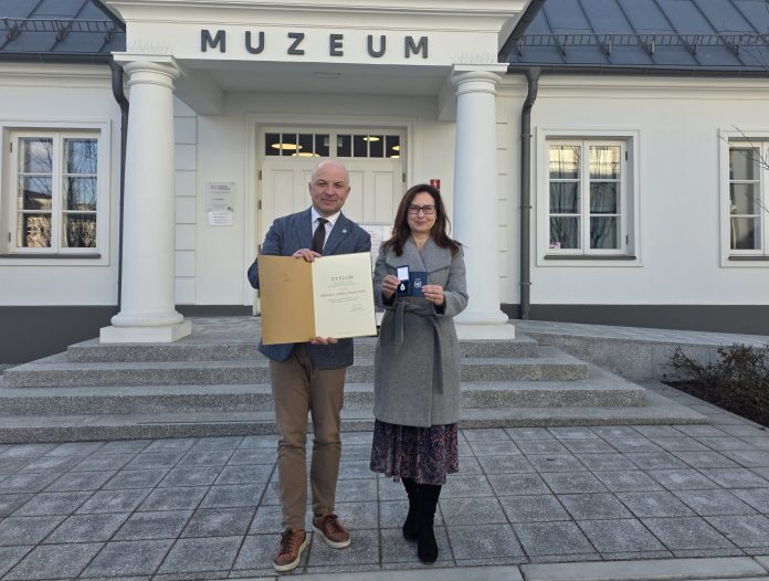 Burmistrz i dyrektor muzeum pozują pod budynkiem muzeum z dyplomem i medalem
